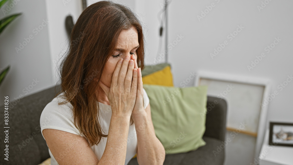 Mature woman in distress with hands covering face sitting on couch at home, conveying emotion of sadness or worry. Mature woman in distress with hands covering face sitting on couch at home, conveying emotion of sadness or worry.