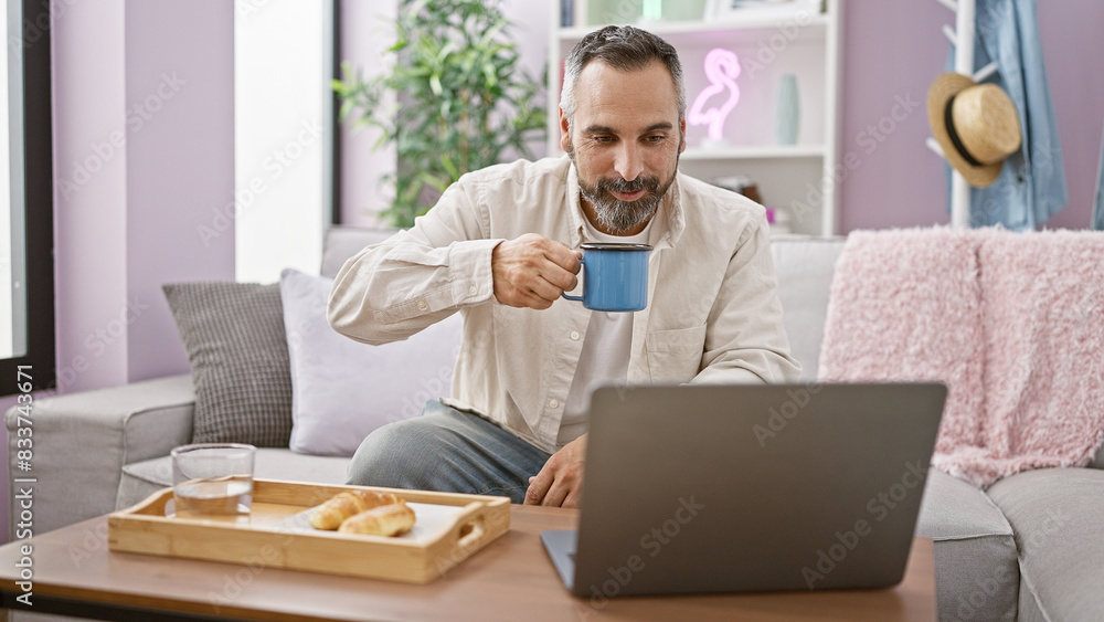 Mature hispanic man with beard using laptop and drinking coffee at home Mature hispanic man with beard using laptop and drinking coffee at home