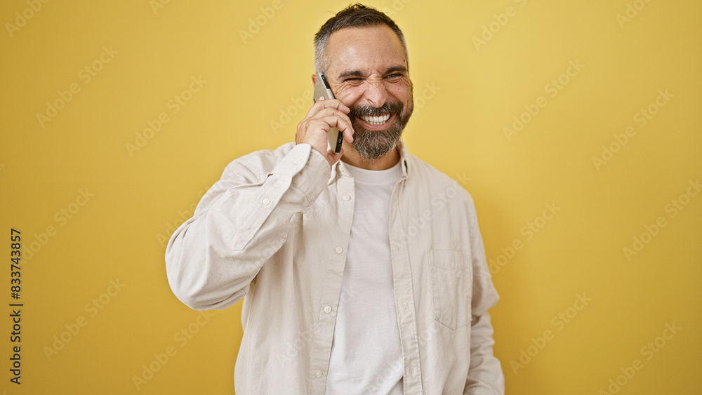 Obraz premium Mature hispanic man with beard laughing on phone against yellow background