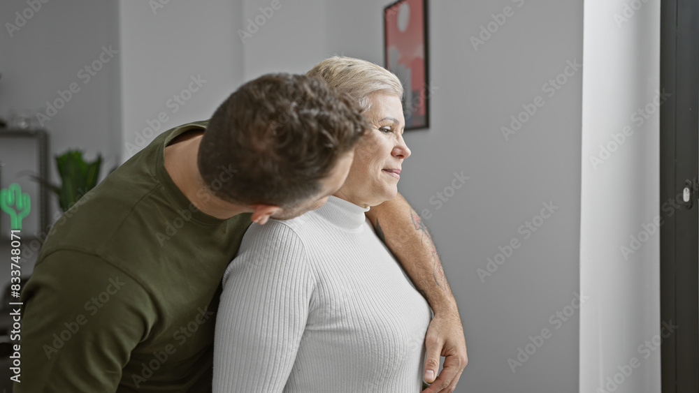 A tender moment as a young man embraces an older woman in a cozy modern living room, conveying family affection. A tender moment as a young man embraces an older woman in a cozy modern living room, conveying family affection.
