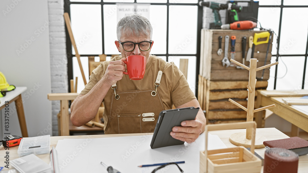 A grey-haired man enjoys a coffee break in a woodworking workshop, browsing on a tablet amidst handcrafted woodwork. A grey-haired man enjoys a coffee break in a woodworking workshop, browsing on a tablet amidst handcrafted woodwork.