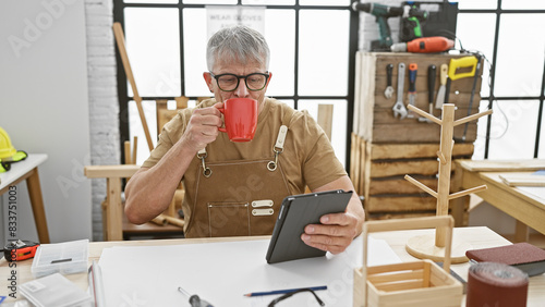 A grey-haired man enjoys a coffee break in a woodworking workshop, browsing on a tablet amidst handcrafted woodwork.