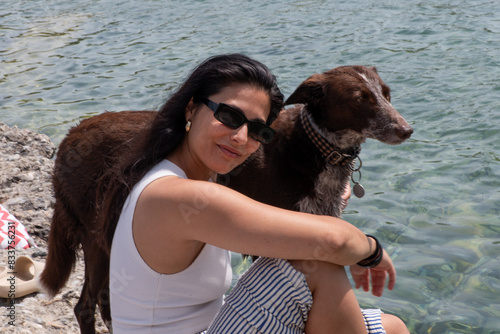 woman with sunglasses, long brown hair, standing next to a dog in the river, wearing striped trousers and a white tank top, looking at the camera