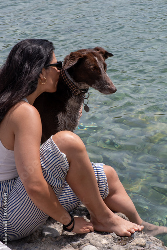 woman with sunglasses long brown hair kissing a dog in the river, wearing striped trousers and a white tank top.