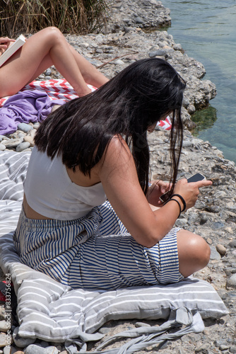 Woman chatting on mobile phone in river landscape, dressed in striped trousers and white tank top, long black hair, in the background legs of a girl sunbathing and reading.