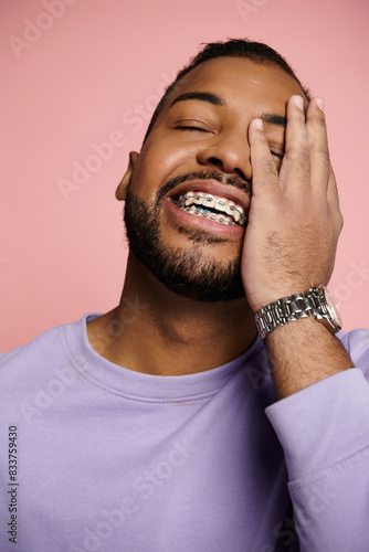 A young African American man smiling brightly, showcasing his braces, against a vibrant pink background.