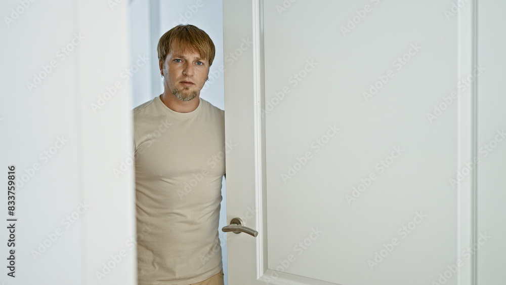 Serious blond man with beard standing indoors by an open door looking at camera Serious blond man with beard standing indoors by an open door looking at camera