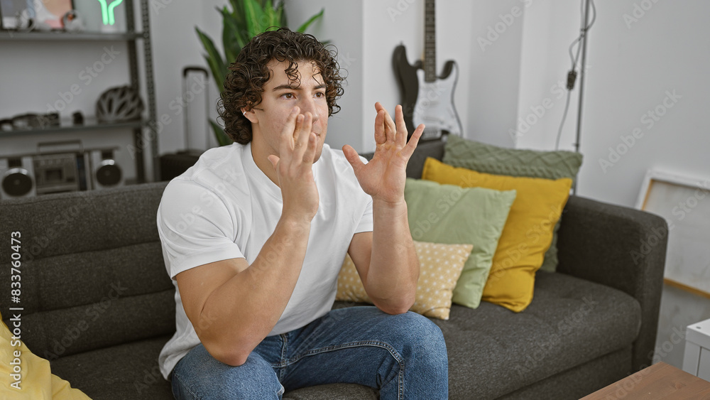 Young hispanic man with curly hair gesturing while sitting on couch in a cozy living room. Young hispanic man with curly hair gesturing while sitting on couch in a cozy living room.