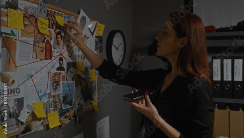 Redhead woman detective analyzing evidence board in office while holding phone