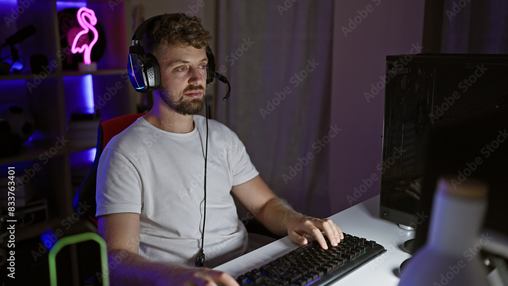 Caucasian bearded young man with blue eyes focusing on his computer in a dimly lit gaming room at night. Caucasian bearded young man with blue eyes focusing on his computer in a dimly lit gaming room at night.