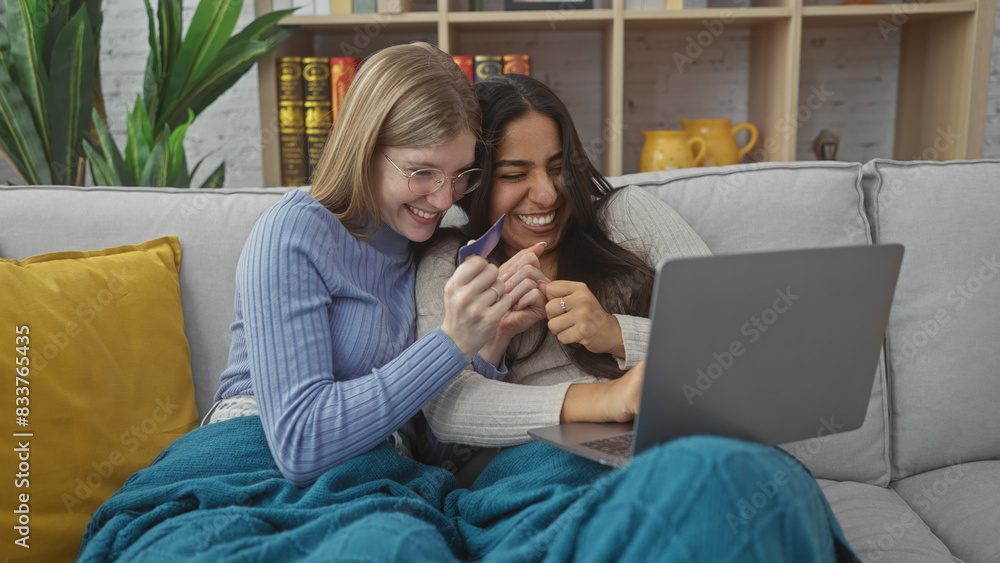 Two women laugh together on a couch while holding a credit card and looking at a laptop in a cozy living room.