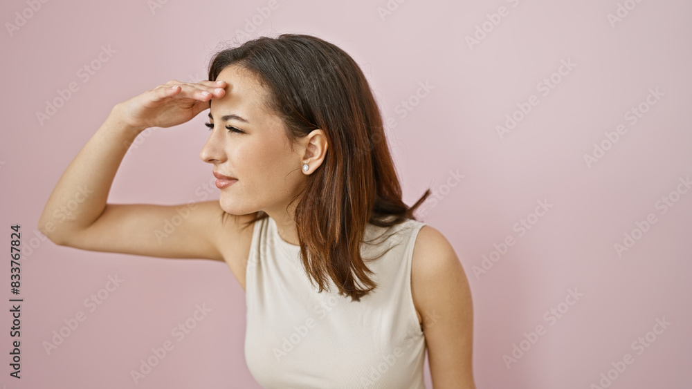 A young hispanic woman in a beige dress poses with a thoughtful expression against a pink wall.