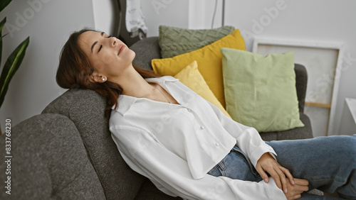 Obraz na plátně Relaxed hispanic woman reclining on a gray sofa at home surrounded by colorful pillows