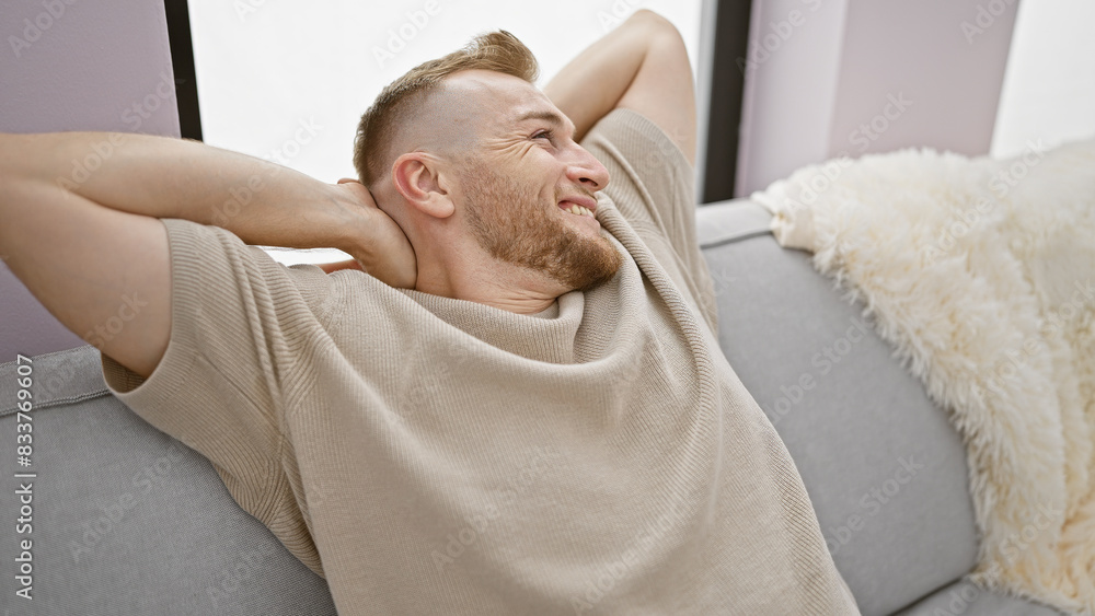 A relaxed young man with a beard sitting on a sofa, enjoying his cozy living room at home. A relaxed young man with a beard sitting on a sofa, enjoying his cozy living room at home.