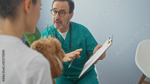Hispanic veterinarian consults with a woman holding a poodle inside a clinic room.