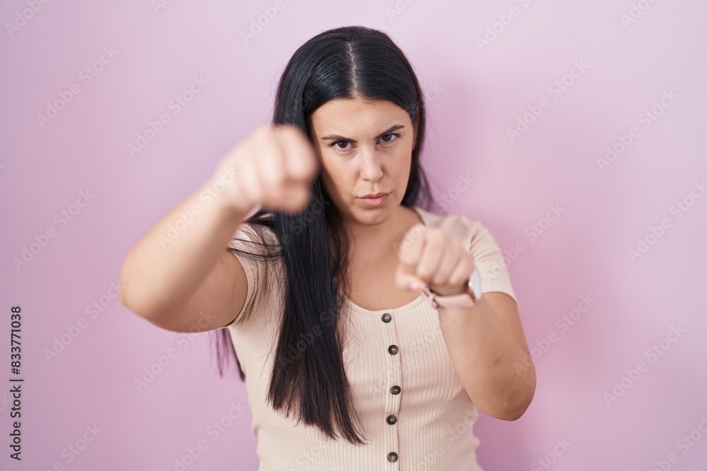 Fototapeta premium Young hispanic woman standing over pink background punching fist to fight, aggressive and angry attack, threat and violence