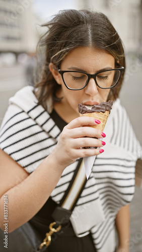 A young attractive hispanic woman enjoys a chocolate ice cream cone on a bustling city street.