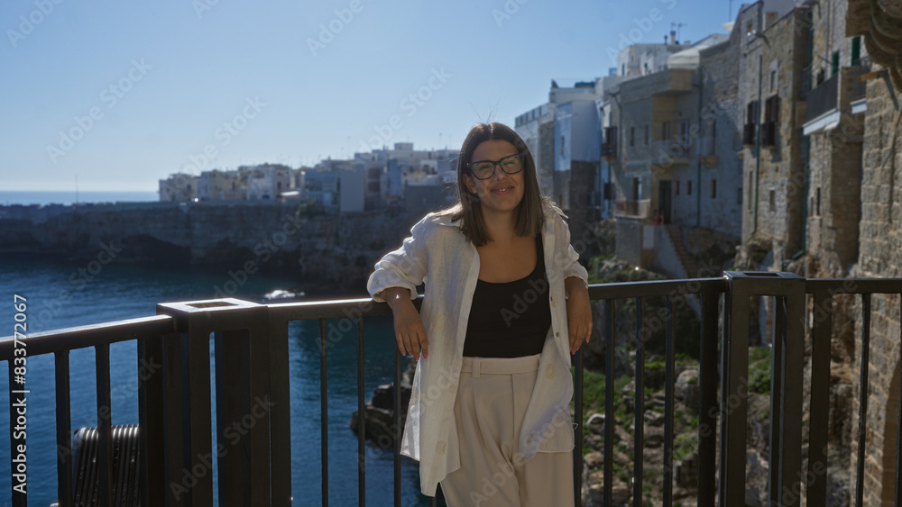 A young, beautiful hispanic woman smiles at the viewpoint in polignano a mare, puglia, italy, overlooking the scenic coastline and picturesque buildings while basking in the sunny weather. A young, beautiful hispanic woman smiles at the viewpoint in polignano a mare, puglia, italy, overlooking the scenic coastline and picturesque buildings while basking in the sunny weather.