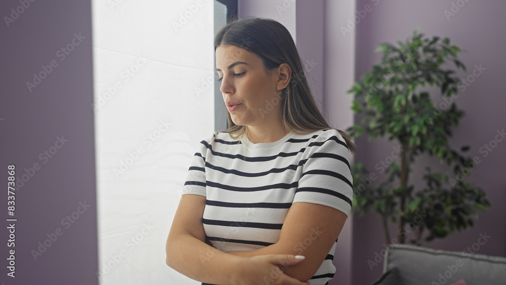 Attractive young hispanic brunette woman with crossed arms stands indoors by a window in a modern living room, appearing contemplative and serene.