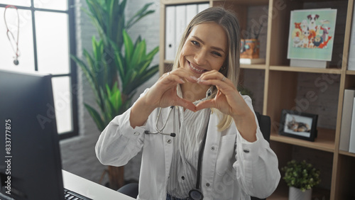 A beautiful young blonde woman in a clinic, wearing a stethoscope, making a heart gesture with her hands, while smiling warmly in an indoor medical workplace. photo