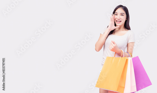 A cheerful young Asian woman in casual attire, holding colorful shopping bags and making an announcement gesture against a white background. The image conveys excitement, enthusiasm, joy of shopping