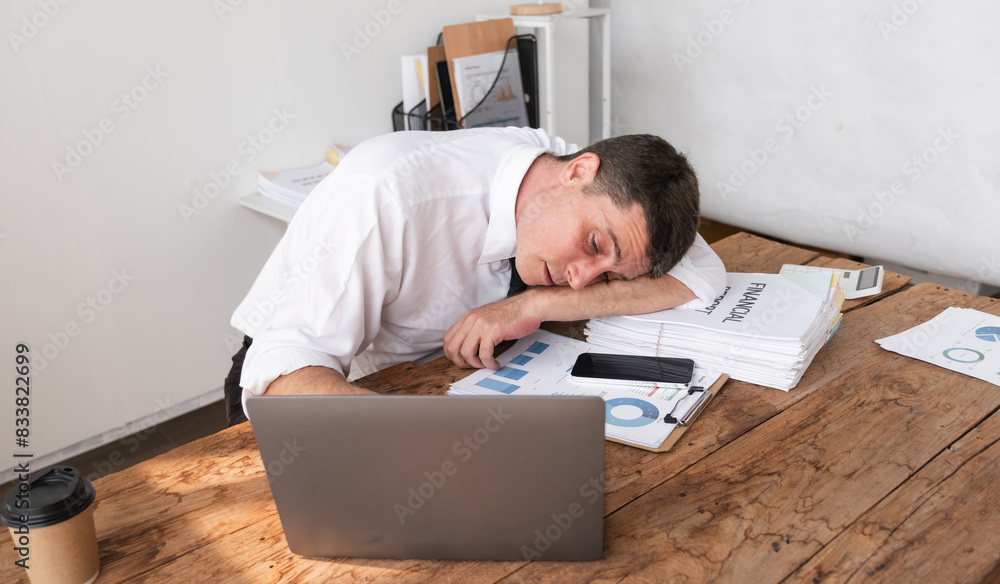 Obraz premium Businessman exhausted and asleep at desk with laptop and financial reports, concept of workplace fatigue and stress