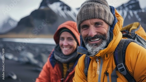Two hikers one with a beard smiling at the camera wearing winter gear and carrying backpacks standing in front of a snow-capped mountain range.