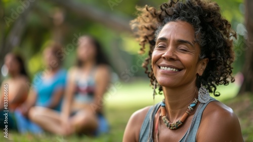 A woman with curly hair smiling at the camera sitting cross-legged on grass with blu rred figures of other people in the background suggesting a peaceful outdoor gathering or event. photo