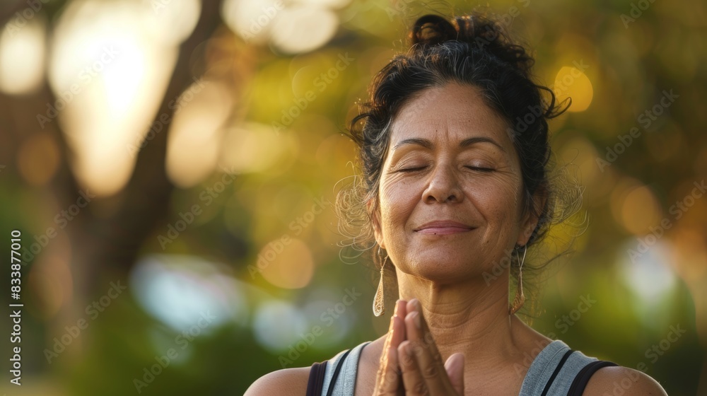 A woman with closed eyes smiling and holding her hands together in a prayerful or meditative pose with a blurred background of trees and sunlight.