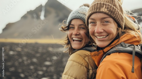 Two women hiking smiling wearing winter gear and carrying backpacks with a mountainous landscape in the background.