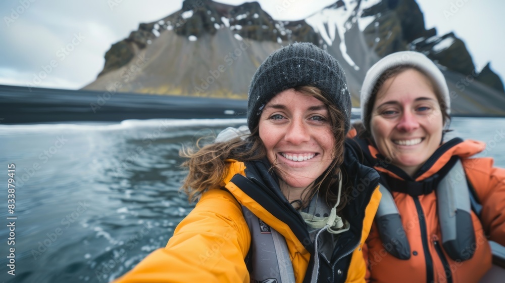 Two women in winter gear, smiling at the camera, on a boat with a mountainous landscape in the background.