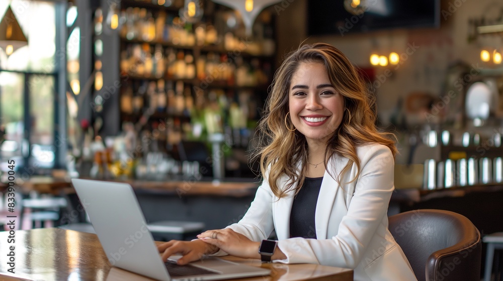 Fototapeta premium Happy young Latina businesswoman in suit working with laptop in a cafe or bar smiling confidently, for business owner, self employment, work from home, manager, small business, remote working concept.
