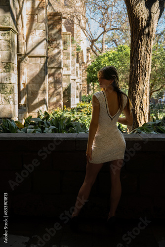 Beautiful young woman in a short, tight white dress posing in profile standing stylishly looking indifferently with a cloister garden illuminated by the evening light. photo