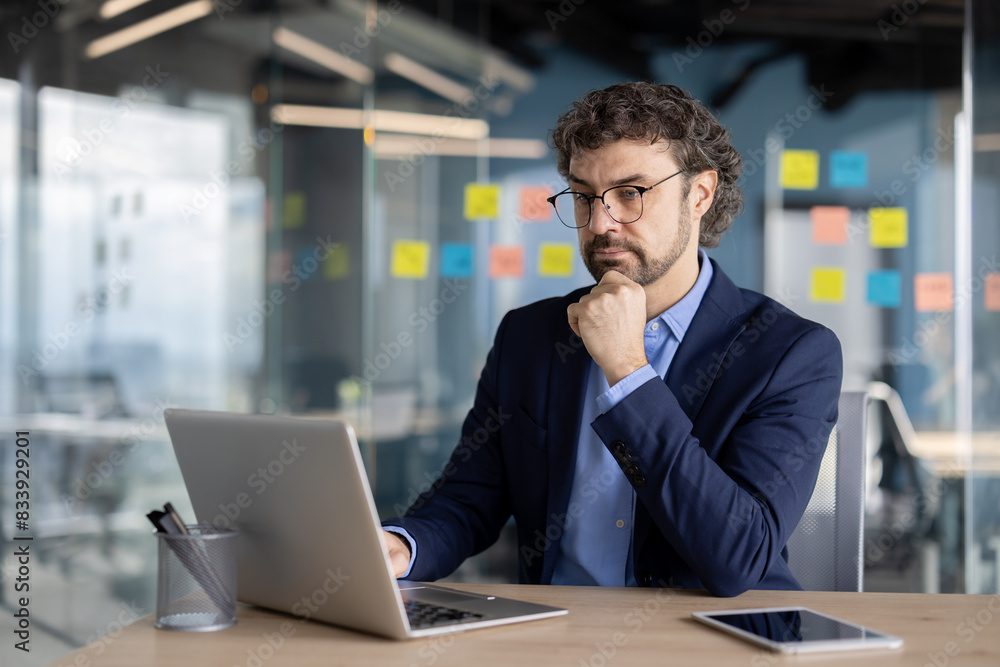 Thinking businessman inside office at workplace, mature experienced man in business suit working with laptop, boss focused, looking at electronic reports and investment proposals. Thinking businessman inside office at workplace, mature experienced man in business suit working with laptop, boss focused, looking at electronic reports and investment proposals.