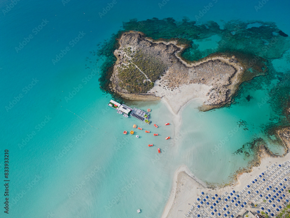 Aerial view of Nissi island and empty beach in Ayia Napa, Cyprus. Calm turquoise Mediterranean sea on a quiet sunny morning. No people. Sandy beach with lines of parasols and watersports business.