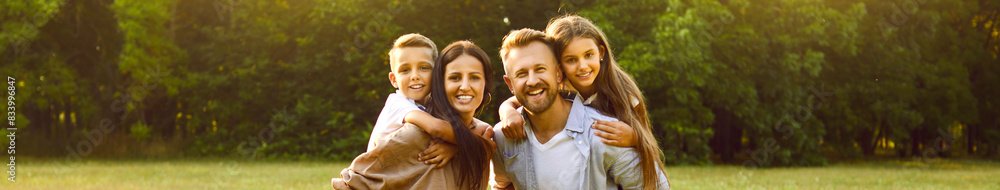 Portrait of happy smiling family of four with son and daughter standing in the summer park and looking cheerful at camera. Parents with two kids walking in nature enjoying sunny day together. Banner.