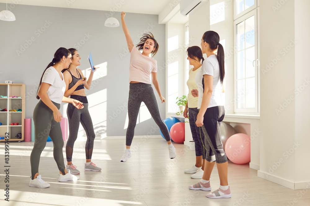 Fototapeta premium Happy female athlete having fun after successful fitness workout at gym. Energetic overjoyed young woman breaks a sports record, celebrates together with friends and jumps up in excitement