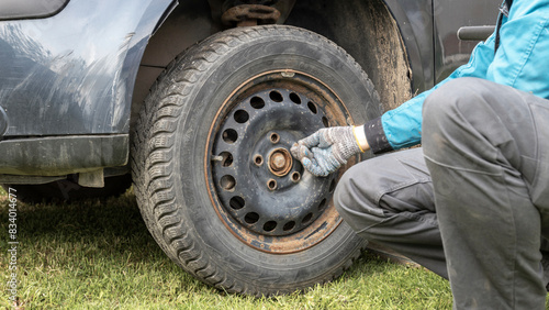 A person is kneeling beside a car, changing a dirty, muddy tire on a sunny day. The car is parked on grass, and the individual is wearing gloves and a jacket