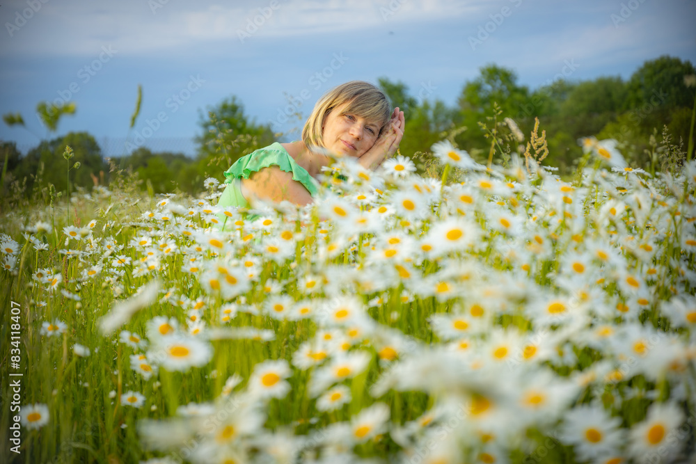 Fototapeta premium A woman in a green shirt rests her head on her hands in a field of blooming white daisies with yellow centers. The background features lush greenery and a clear sky, creating a serene and peaceful sce