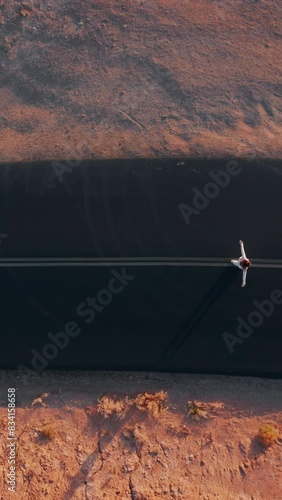 Vertical Screen: Aerial view captures the contrast between nature and humanmade infrastructure as a lone girl walks on a straight, black road in a desert, symbolizing solitude and exploration amidst photo