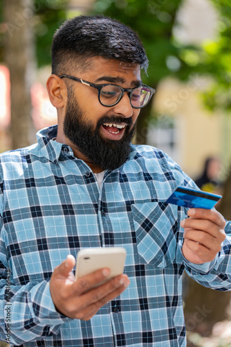 Shocked Indian man trying to pay online shopping with smartphone blocked credit card. Annoyed bearded Hispanic tourist on street buying bank refuse problem unsuccessful payment lack of money balance