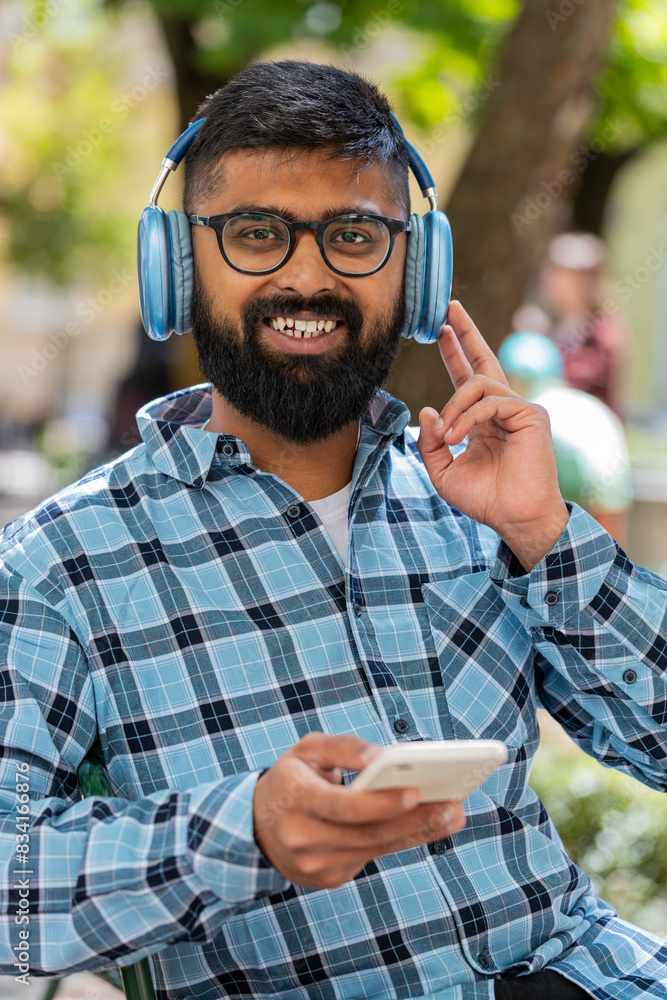Happy overjoyed Indian bearded man dancing in wireless headphones listening favorite energetic music, celebrating victory win outdoors. Hispanic guy on urban city street. Town lifestyles. Vertical Happy overjoyed Indian bearded man dancing in wireless headphones listening favorite energetic music, celebrating victory win outdoors. Hispanic guy on urban city street. Town lifestyles. Vertical