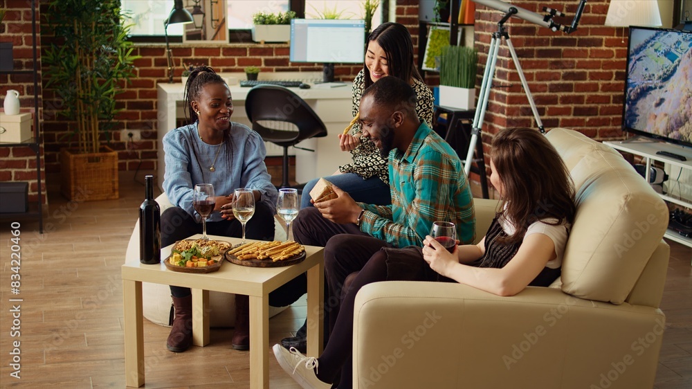 Multiethnic mates invited to birthday party offering presents to cheerful African american man impressed by nice gesture. Guests surprising their friend with thoughtful present and enjoying appetizers Multiethnic mates invited to birthday party offering presents to cheerful African american man impressed by nice gesture. Guests surprising their friend with thoughtful present and enjoying appetizers