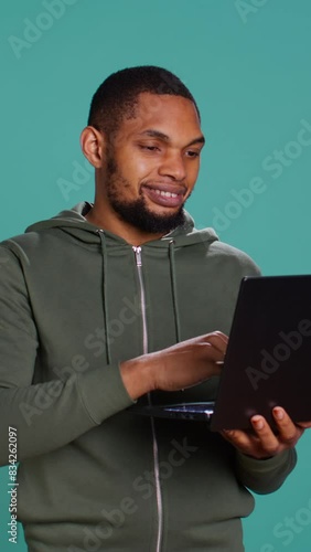 Vertical video Smiling man scrolling on laptop, using internet and credit card to pay for products. African american person happily browsing online shopping websites, isolated over studio background photo