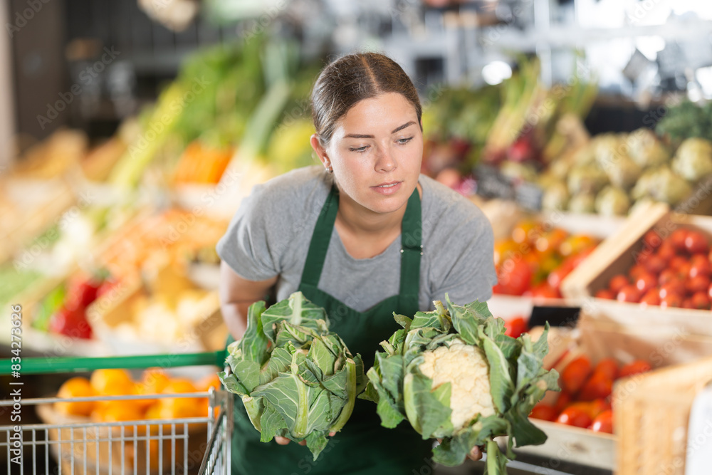 Enthusiastic young female grocery worker in green apron arranging fresh organic cauliflower on vibrant vegetable display in farm produce section
