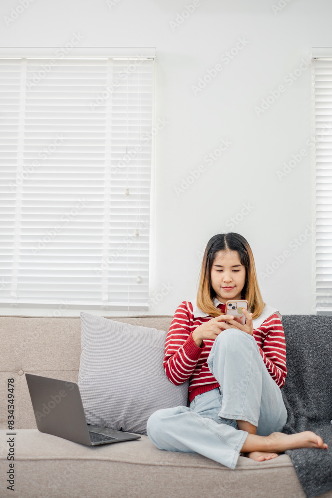 A woman is sitting on a couch with a laptop and a cell phone in her hands. She is wearing a red and white striped shirt and blue jeans. Concept of relaxation and leisure A woman is sitting on a couch with a laptop and a cell phone in her hands. She is wearing a red and white striped shirt and blue jeans. Concept of relaxation and leisure