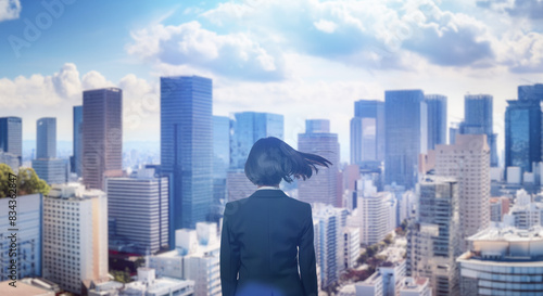 日本の街・都市・朝日を受けたオフィスビル群を前に佇み風を受けて髪をなびかせるスーツを着た会社員の女性の出勤 ビジネス・はじまり・決意・目標・挑戦のイメージ