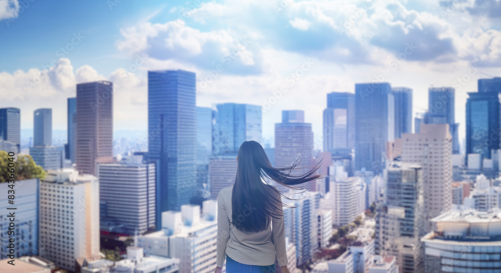 Obraz premium 日本の街・都市・朝日を受けたオフィスビル群を前に佇み風を受けて髪をなびかせる会社員の女性の出勤 ビジネス・はじまり・決意・目標・挑戦のイメージ