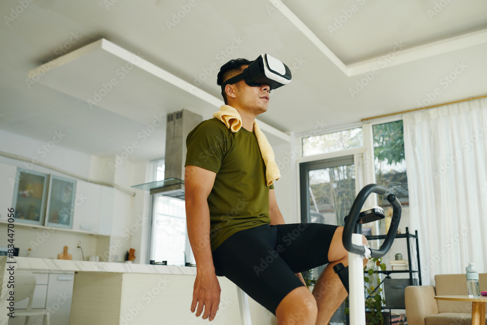 © DragonImages - A man sitting on a stationary bike, resting after a virtual reality exercise session in a spacious, well-lit living room