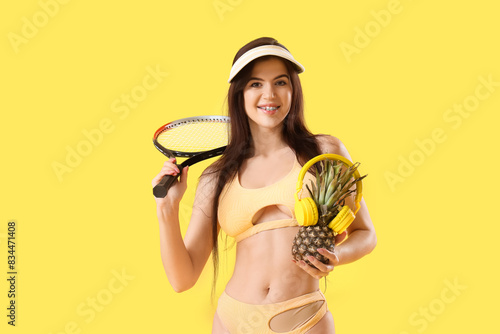 Portrait of pretty young woman in swimsuit with pineapple, headphones and tennis racquet on yellow background